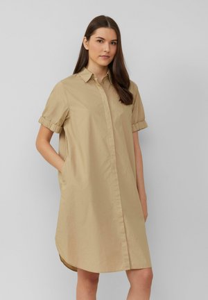 Abito camicia beige con maniche corte arrotolate, chiusura frontale con bottoni e tasche laterali. Tessuto liscio, lunghezza al ginocchio con orlo curvo.