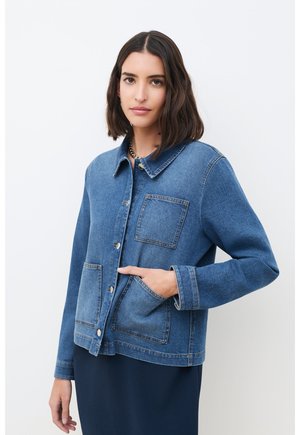 Femme aux cheveux sombres mi-longs portant une veste en jean bleue avec trois poches frontales sur une jupe sombre, debout devant un fond blanc.
