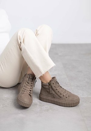 Zapatillas altas marrones de ante texturizado, con cordones en la parte frontal, cremallera lateral y suela gruesa estriada; usadas con pantalones color crema.
