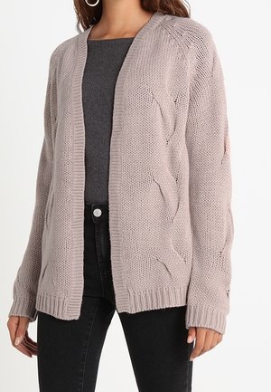 Femme portant un cardigan épais tricoté mauve clair avec des motifs torsadés sur un top gris foncé et un jean noir, debout, les bras détendus.