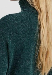 Pull col roulé en tricot texturé vert foncé avec un col haut ; cheveux blonds longs et raides partiellement visibles.
