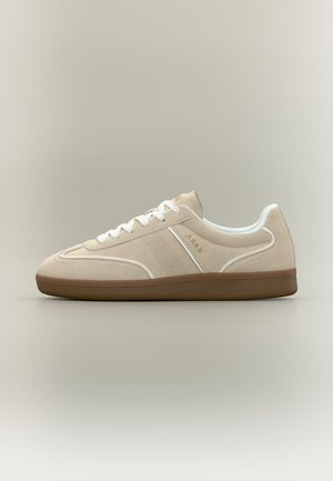 Bézs színű szarvasbőr sneaker fehér fűzőkkel, fehér szegéllyel, perforált oldalsó panellel, gumitalpú talppal és „ARKK” márkajelzéssel az oldalon.