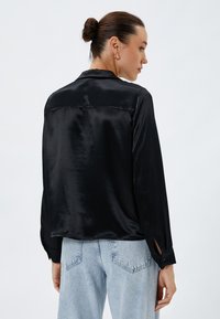 Zwarte satijnen blouse met een iets glanzende textuur, lange mouwen en detaillering op de schoudernaden, gecombineerd met lichtblauwe jeans.
