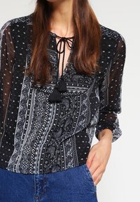 Blusa de estampado en blanco y negro con cuello de lazo, acentos de borlas y mangas transparentes, combinada con jeans de mezclilla azul.