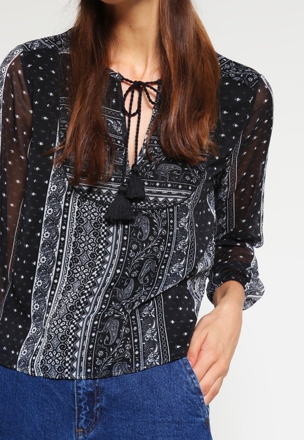 Blusa de estampado en blanco y negro con cuello de lazo, acentos de borlas y mangas transparentes, combinada con jeans de mezclilla azul.