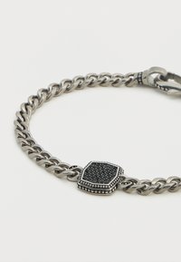 Βραχιόλι από μεταλλικό υλικό ασημί απόχρωσης με χοντρή αλυσίδα, κεντρικό διακοσμητικό με υφή μαύρης πέτρας και λεπτομερείς άκρες.