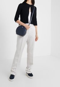 Navy runde Umhängetasche aus glattem Leder, silberne Hardware, verstellbarer Riemen, kombiniert mit einer schwarzen kurzärmligen Strickjacke und weißen Hosen.