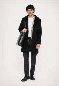 Abrigo de lana negra con capucha, capa interior beige, pantalones grises y zapatos negros. Lleva un bolso estructurado negro.