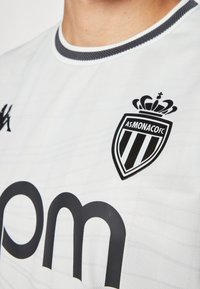 Witte voetbalshirt met een gestructureerde afwerking, voorzien van een zwart logo met een kroon en verticale strepen, en een donkere ronde kraag.