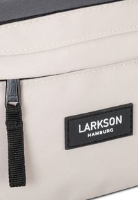 Világos bézs táska sötét szürke részletekkel, textil mintázattal, cipzáras díszítéssel és egy fekete gumi márkacímkével, amelyen a "LARKSON HAMBURG" felirat olvasható.