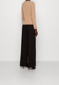 Beige Strickpullover mit langen Ärmeln, kombiniert mit hoch taillierten schwarzen, weit geschnittenen Hosen und Stilettos, von hinten betrachtet.