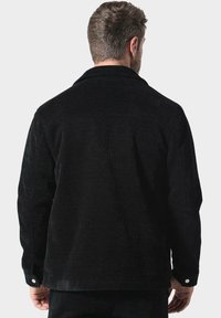 Muž s krátkými hnědými vlasy, oblečený v černé texturované dlouhé bundě, zobrazený zezadu na jednolitém světlém pozadí.