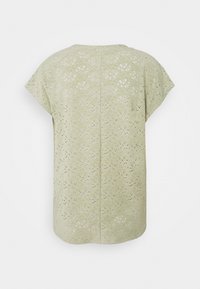 Blouse vert clair à manches courtes avec motif en dentelle florale à œillets et ourlet arrondi visible de dos sur un fond uni.