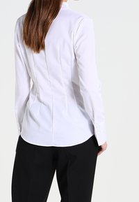 Weißes passgenaues Hemd mit Stehkragen, langen Ärmeln, Rückenfalten und Knopf-Details an den Ärmelbündchen. Glatte Textur. Wird mit schwarzen Hosen getragen.