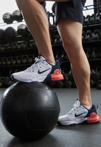 Białe i granatowe buty sportowe z teksturowaną cholewką i wyraźnymi czerwonymi akcentami, umieszczone na czarnej piłce lekarskiej w tle siłowni.