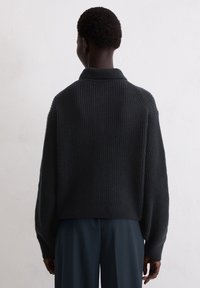 Pull en maille gris foncé avec une texture côtelée, col et épaules tombantes. Longueur courte, porté avec un pantalon foncé.