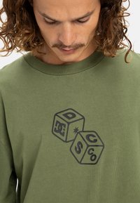 Olive green cotton t-shirt featuring black graphic print of two dice with "D" and "S" on them, round neckline, and relaxed fit.