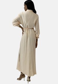 Largo vestido de melocotón pálido con un patrón texturizado, cintura ceñida, mangas anchas y abertura lateral. Llevado con tacones de color claro.