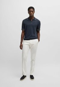 Granatowa koszulka polo z zamkiem i białymi wykończeniami, zestawiona z białymi spodniami i czarnymi butami wsuwanymi. Prosta, nowoczesna sylwetka.