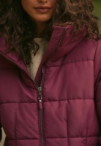 Veste doudoune bordeaux avec un design matelassé, col montant et détail de fermeture éclair à l'avant, portée sur un pull en tricot de couleur claire.