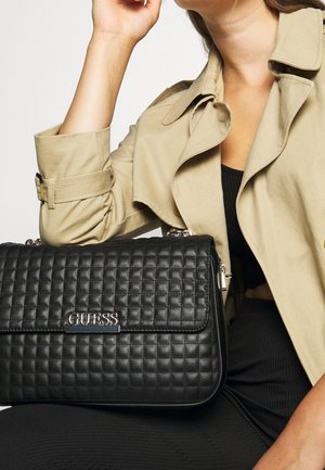 Femme portant un trench beige et un haut noir, tenant un sac à main matelassé noir Guess avec logo argenté sur ses genoux.