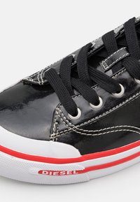 Sneaker in pelle verniciata nera con lacci neri, occhielli in argento e accenti rossi sulla suola. Presenta una punta in gomma bianca e cuciture dettagliate.