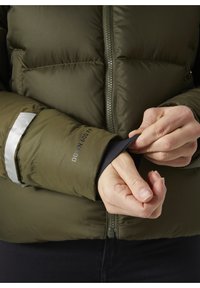 Olijfgroene pufferjack met een geribbelde polsband, voorzien van reflecterende details en een rits aan de voorkant. De tekst op de mouw geeft het type isolatie aan.