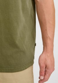 Olivenfarget bomulls-t-skjorte med en lett teksturert overflate; har et merke på siden og er parret med brune shorts.