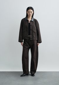 Brown patterned jacket with a contrast black collar, paired with wide brown pants and black pointed shoes. Plain background.