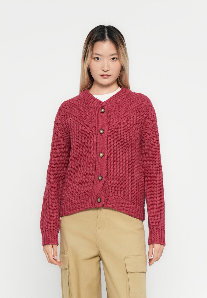 Γυναίκα φορεμένη με μια υφασμάτινη ροζ φούξια καρδιολίνου με κουμπιά και μπεζ παντελόνι cargo, στέκεται σε έναν απλό λευκό φόντο.