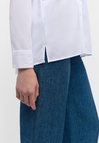 Weißes Langarmhemd mit einem scharfen Kragen, mit einem seitlichen Schlitz am Saum, kombiniert mit dunkelblauen strukturierten Jeanshosen.