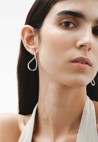 Pendientes de plata con forma de gota, con una superficie lisa y reflectante, colgando de la oreja de la modelo, que muestran un diseño minimalista.