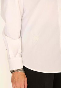 Chemise blanche à boutons, avec une texture douce, ornée d'un monogramme brodé, manches longues et ourlet arrondi. Bracelets en argent au poignet.