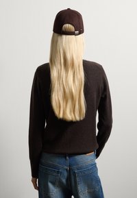 Cappello marrone con cinturino regolabile, indossato con un maglione scuro e jeans blu. Capelli biondi lunghi e mossi visibili, rivolti lontano dalla telecamera.