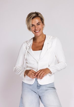 Smiling woman with short blonde hair wearing a white buttoned jacket over a top and light blue jeans, standing against a plain background.