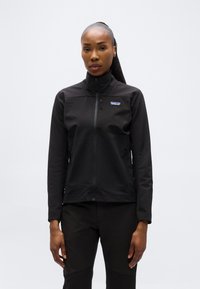 Γυναίκα με μαλλιά πιασμένα πίσω, φορώντας μαύρο μπουφάν Patagonia με φερμουάρ και μαύρο παντελόνι, που στέκεται μπροστά σε απλό φωτεινό φόντο.