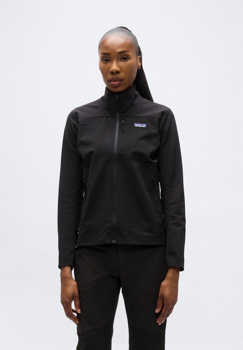 Γυναίκα με μαλλιά πιασμένα πίσω, φορώντας μαύρο μπουφάν Patagonia με φερμουάρ και μαύρο παντελόνι, που στέκεται μπροστά σε απλό φωτεινό φόντο.