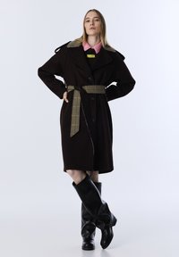 Vrouw in een donkerbruine, ceintuur-jas met een geruite kraag en ceintuur, roze shirt eronder en zwarte kniehoge laarzen, staand tegen een witte achtergrond.