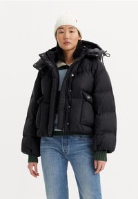 Black puffer jacket with a cropped design, large hood, snap buttons, and front pockets. Worn over a blue and green striped sweater.