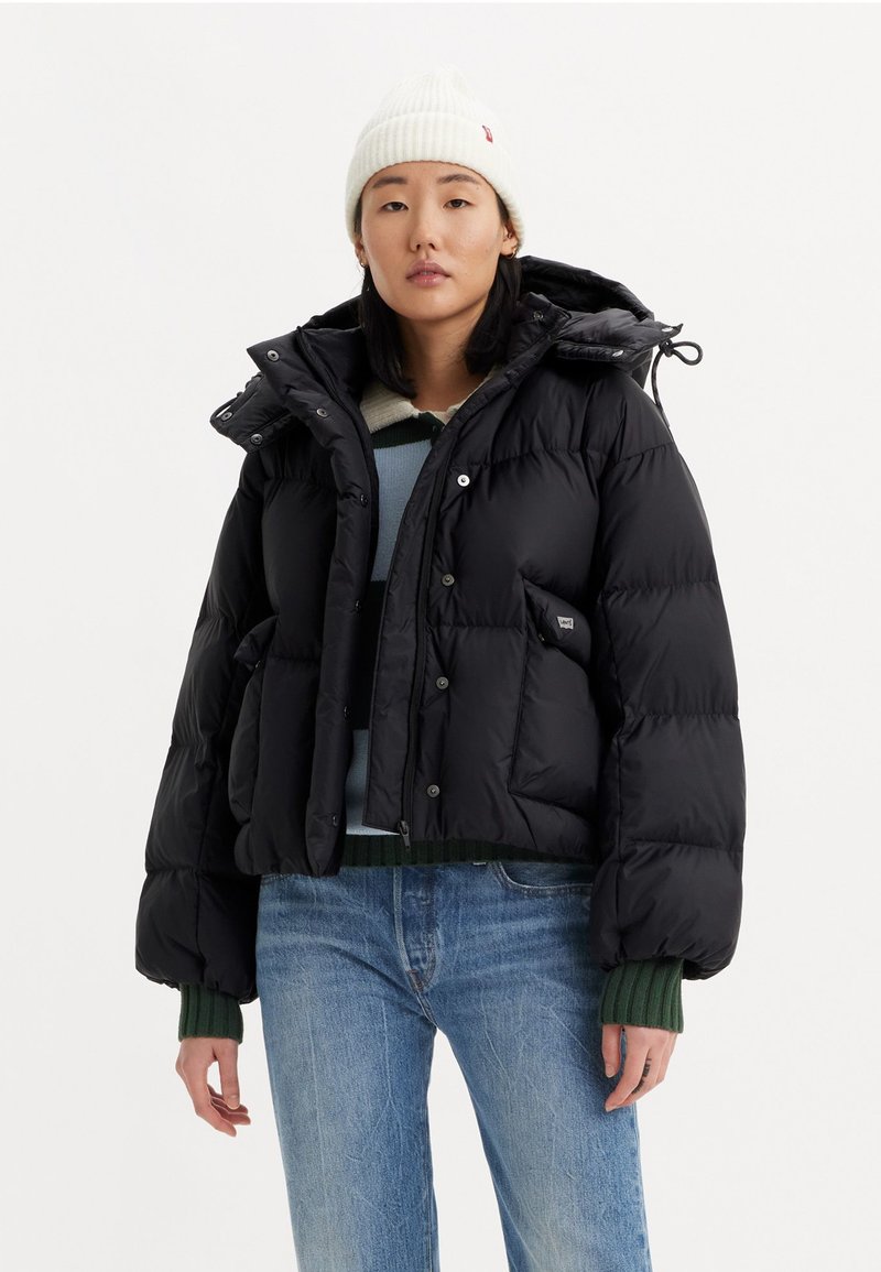 Black puffer jacket with a cropped design, large hood, snap buttons, and front pockets. Worn over a blue and green striped sweater.