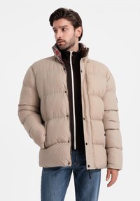 Beige puffjacka med horisontell sömnad, hög krage och rutigt foder. Har tryckknappar och sidofickor. Bärs över en mörk zip-up.