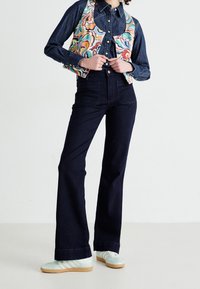 Denim-väst med färgglada abstrakta mönster över en mörkblå denimskjorta och högmidjade mörka jeans, ihop med ljusgröna sneakers.