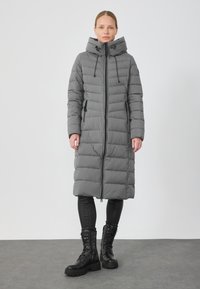 Szürke párnázott hosszú kabát, magas gallérral, állítható kapucnival, elől cipzáras záródással és két oldalzsebből áll. Fekete leggings és csizmák fölött viselve.