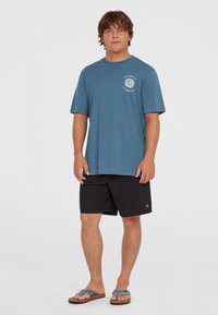 Camiseta azul de manga corta con un gráfico de sol blanco en el lado izquierdo del pecho, combinada con pantalones cortos negros y chanclas.