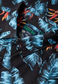 Camisa de manga corta en tela negra con vibrantes hojas azules y acentos florales rojos, con cuello y detalles de botones negros.