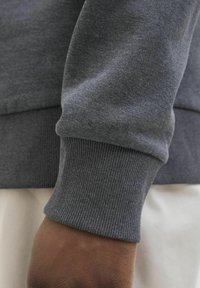 Dunkelhäutige Hand trägt ein dunkelgraues Sweatshirt mit geripptem Bündchen, ruht auf weißem Stoff.