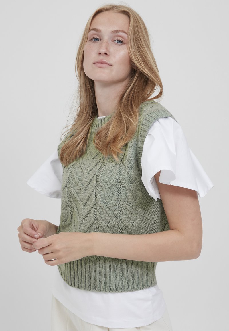 Pulover tricotat de culoare verde deschis, cu un design texturat în cablu, purtat peste un top alb cu mânecă scurtă. Bază cu dantelă și guler rotund.