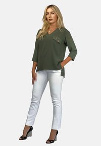 Oliwkowa bluzka z dekoltem w kształcie litery V i kieszonką na piersi, w połączeniu z białymi, dopasowanymi spodniami i czarnymi szpilkami z wzorzystym detalem.