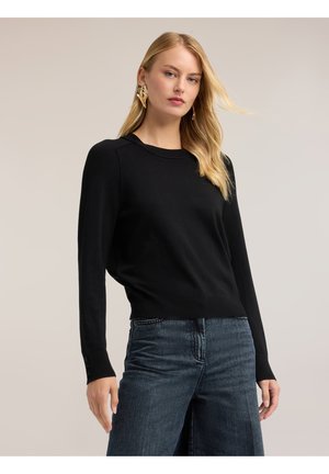 Kvinde med langt blondt hår iført øreringe formet som guldløvblade, en sort langærmet top og mørkeblå jeans mod en ensfarvet baggrund.