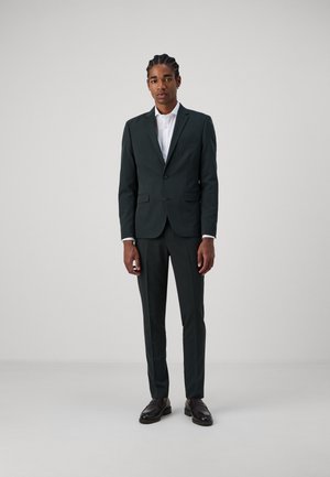 Dark green suit with a fitted blazer and straight-leg trousers. White shirt underneath, paired with black loafers. Minimalist design.